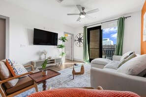 A modern living room with a flat-screen TV, a ceiling fan, a large window with curtains, and a view of the city. - Midtown. Mins to Broadway. Parking. Pool. Gym (Nashville)