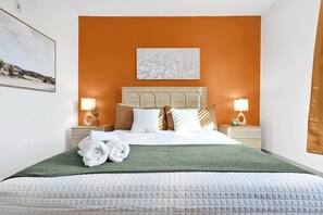 Een hotelkamer met een bed, kussens, een hoofdbord, twee nachtkastjes met lampen, een schilderij aan de muur en een raam met gordijnen.