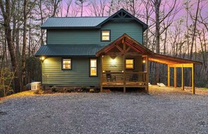 A two-story cabin with a porch and a covered area, surrounded by trees.