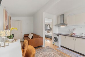 Ein kompakter Wohnraum mit Küchenzeile, einer Couch, einer Waschmaschine und einem Bett im Hintergrund.