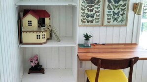 Une pièce avec une table à manger en bois, une chaise jaune, une petite plante en pot, une maquette de maison posée sur une étagère et des motifs de papillons décorant le mur.