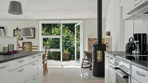 Une cuisine moderne dotée d’un poêle à bois central, de meubles blancs et d’une grande porte-fenêtre coulissante donnant sur un espace patio.