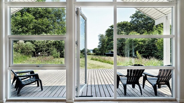 On peut admirer, à travers de grandes fenêtres, une terrasse extérieure dotée d’un plancher en bois, de deux chaises noires et d’un banc.