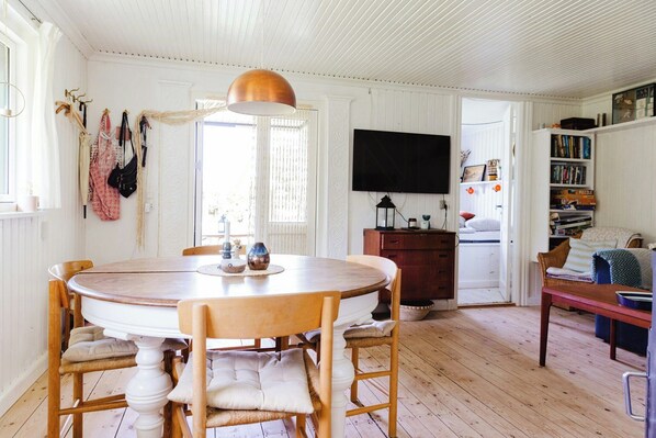 A dining area with a round wooden table, chairs, a pendant light, and a television mounted on the wall. - Romantic island cottage in cosy style near beach (Maribo)