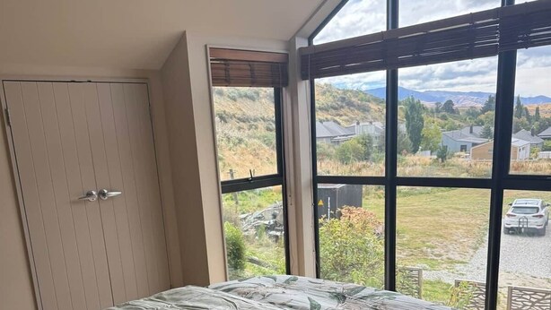 A bedroom with a large window, a bed with floral bedding, and a wardrobe. - Alpine Luxe Cardrona (Cardrona)