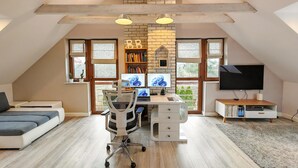 A loft-style bedroom with a sloped ceiling, a desk with two monitors, a TV, and a bookshelf.