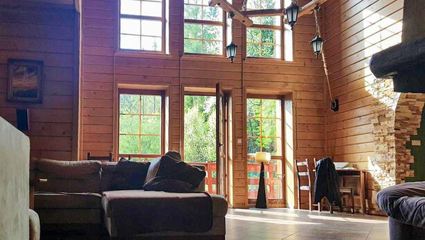 Interior de uma cabana de madeira com um sofá, uma mesa e vista para o exterior através de grandes janelas.