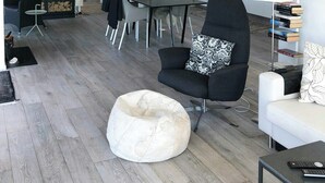 A modern living room with a black armchair, a white bean bag, a dining area with a table and chairs, and a kitchen area with a white fridge and shelves.