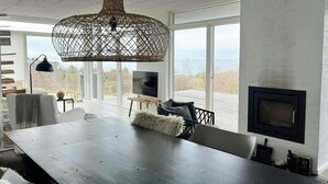 A modern dining room with a dark wooden table, white chairs, a woven pendant light, and a large window offering a view of the outdoors.