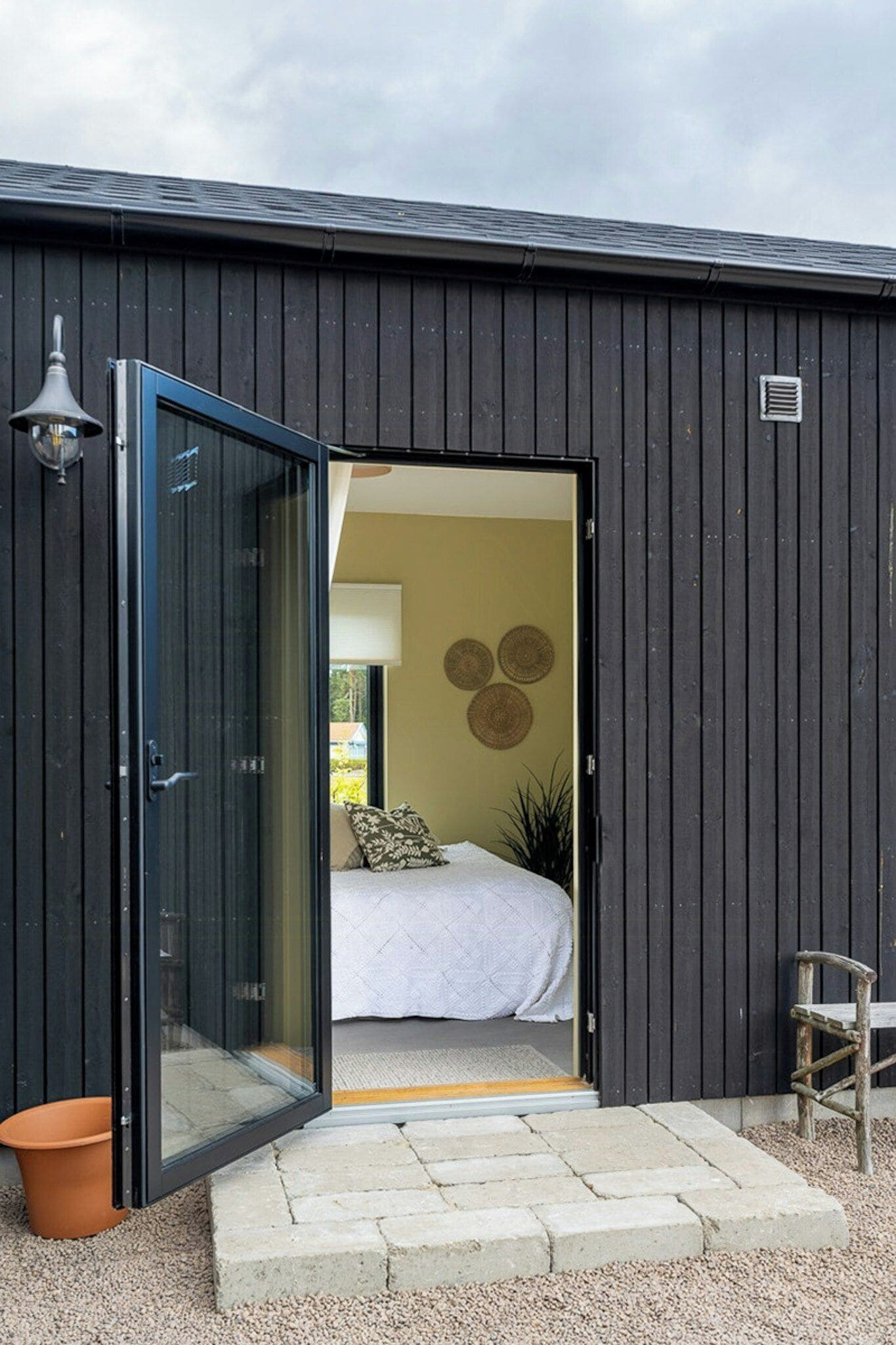 A modern building with a large glass door leading to a room with a bed, a wall-mounted light fixture, and a small potted plant.