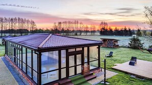 A glass-enclosed building with a tiled roof, a wooden deck, and a grassy area with a view of a body of water and trees. - Awesome home in Darlowo with sauna (Darlowo)