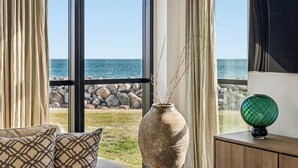 Quarto com uma janela grande com vista para uma costa rochosa, um vaso com galhos e um objeto de vidro verde sobre um armário de madeira.