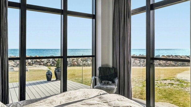 Quarto com janelas amplas, uma cama, uma cadeira e vista para a praia e oceano.