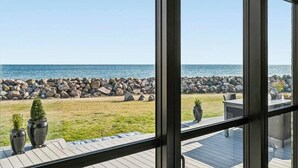 Vista pela janela de um terraço com parede de pedra e plantas, com vista para uma praia e oceano.