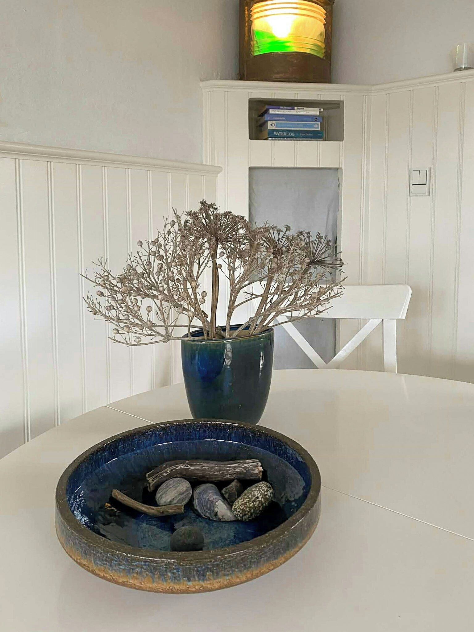 A white table with a blue vase containing dried branches and a bowl of pebbles.