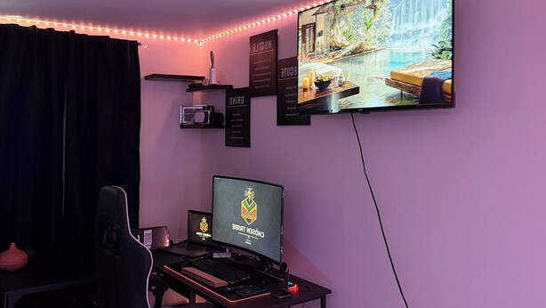 Een kamer met een bureau, stoel, computer en een aan de muur bevestigde televisie waarop een waterval te zien is.