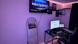 Een kamer met een televisie waarop 'NIGERIAN PRAISE MEDLEY' wordt afgespeeld, een bureau met een laptop, een stoel en een spiegel.