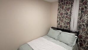 Een enkel bed met een geruit beddegoed en kussens, een gordijn met bloemenprint en een inbouw plafondlamp.