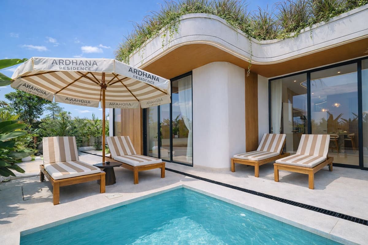 Zona junto a la piscina con tres sillas reclinables y una sombrilla con la etiqueta 'ARDHANA RESIDENCE'.