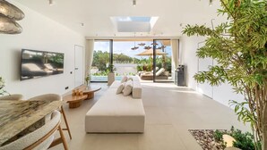 Ein modernes Wohnzimmer mit einer Couch, einem Esstisch und einem Fernseher. Ein großes Fenster bietet Blick auf viel Grün.