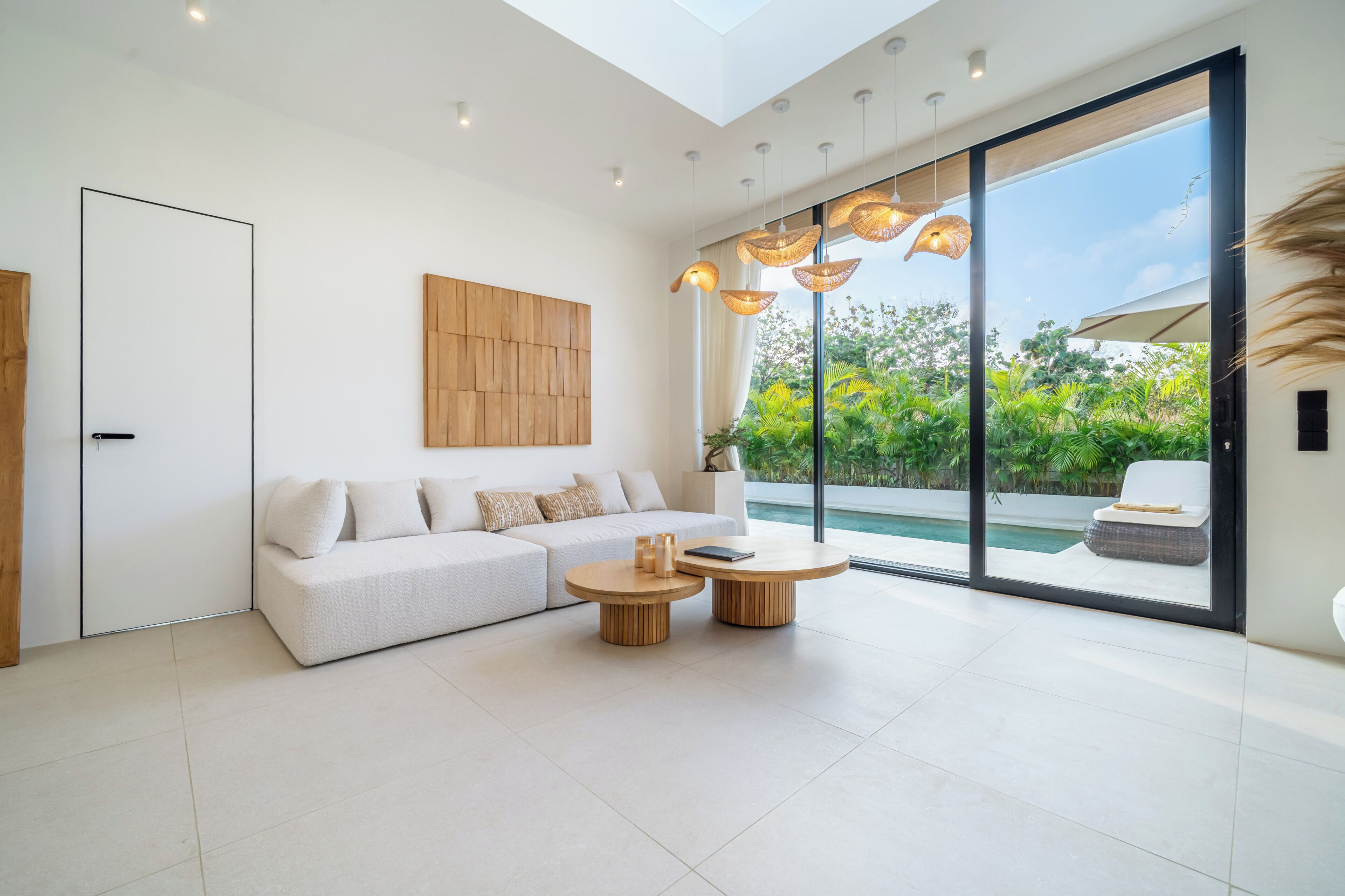 A modern living room with a white sofa, two round wooden tables, and a large sliding glass door leading to a pool.