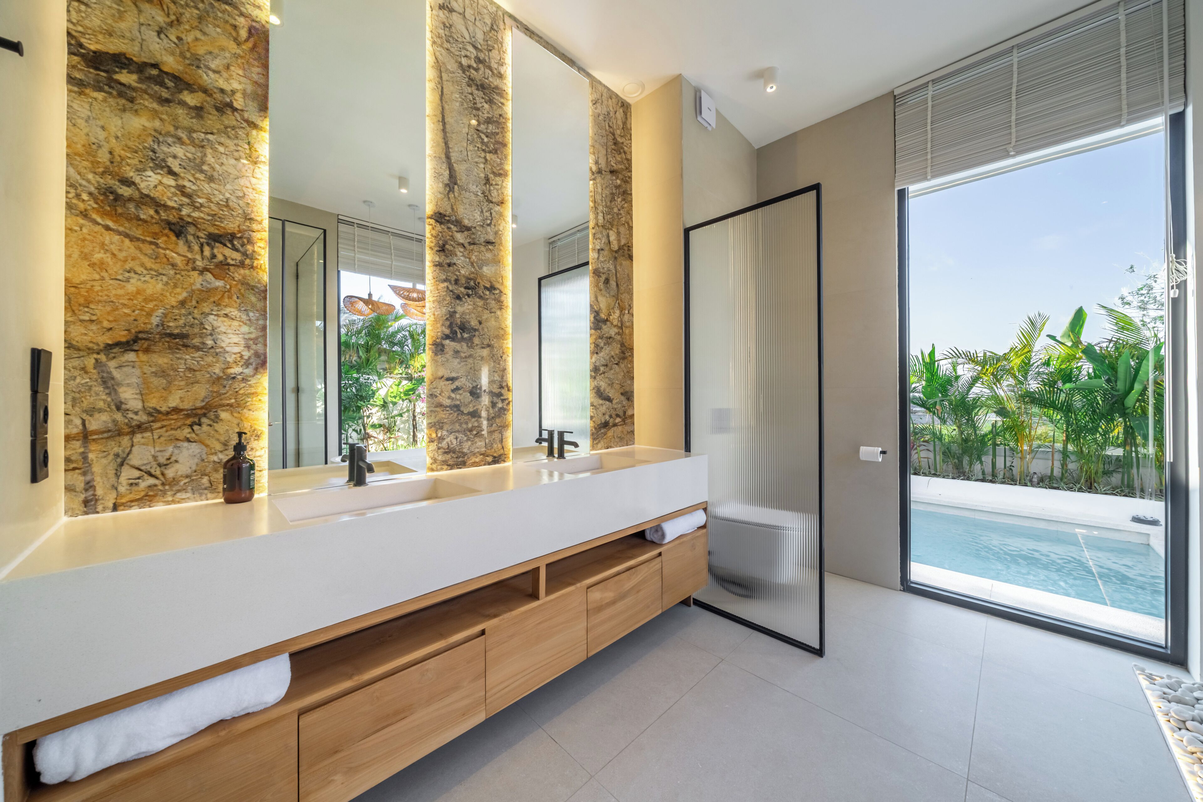 A modern bathroom with a marble wall, large mirror, and a view of a swimming pool.