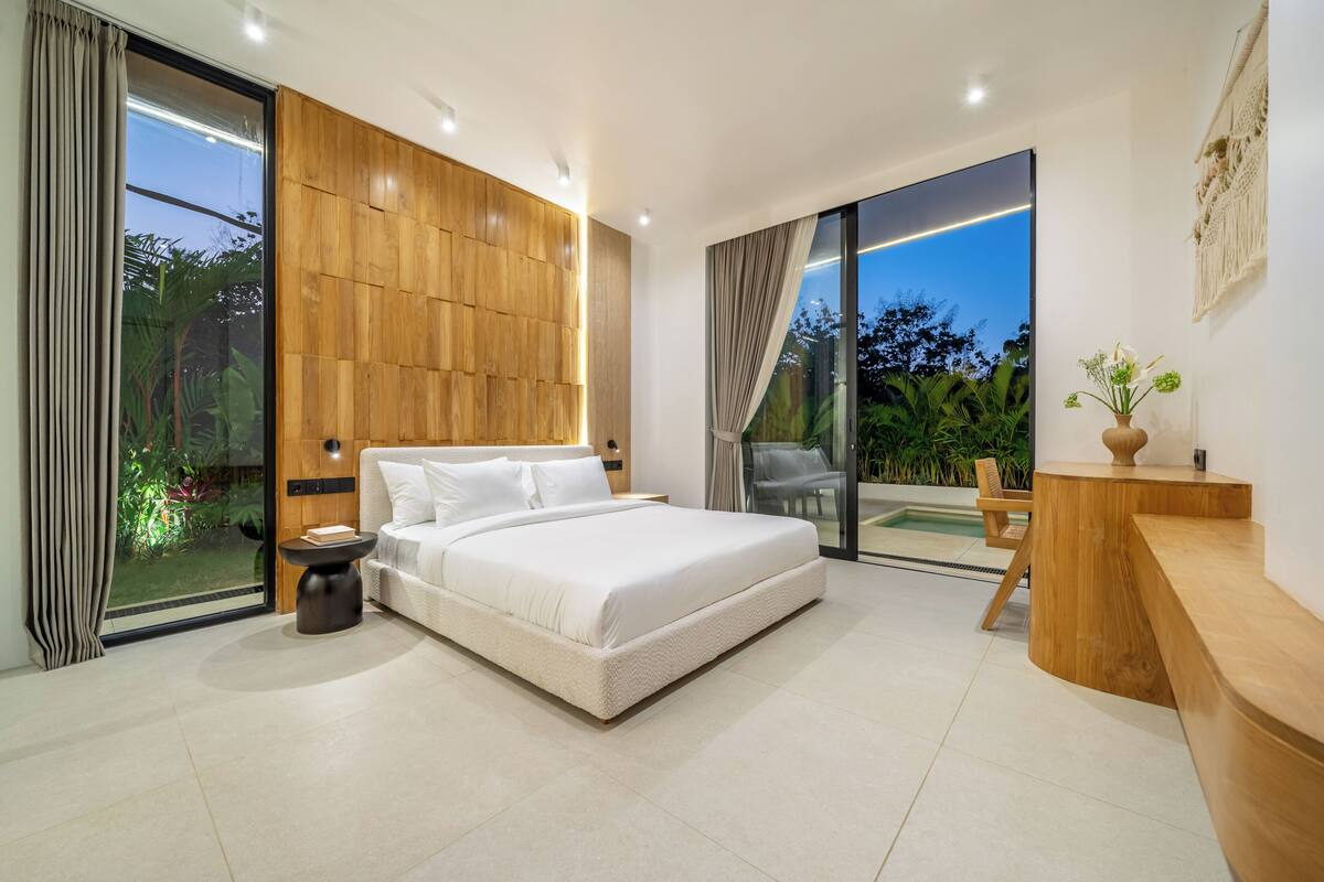 Un dormitorio moderno con una cama grande, un panel decorativo de madera, una puerta de vidrio que da a un balcón y una mesita de noche pequeña.