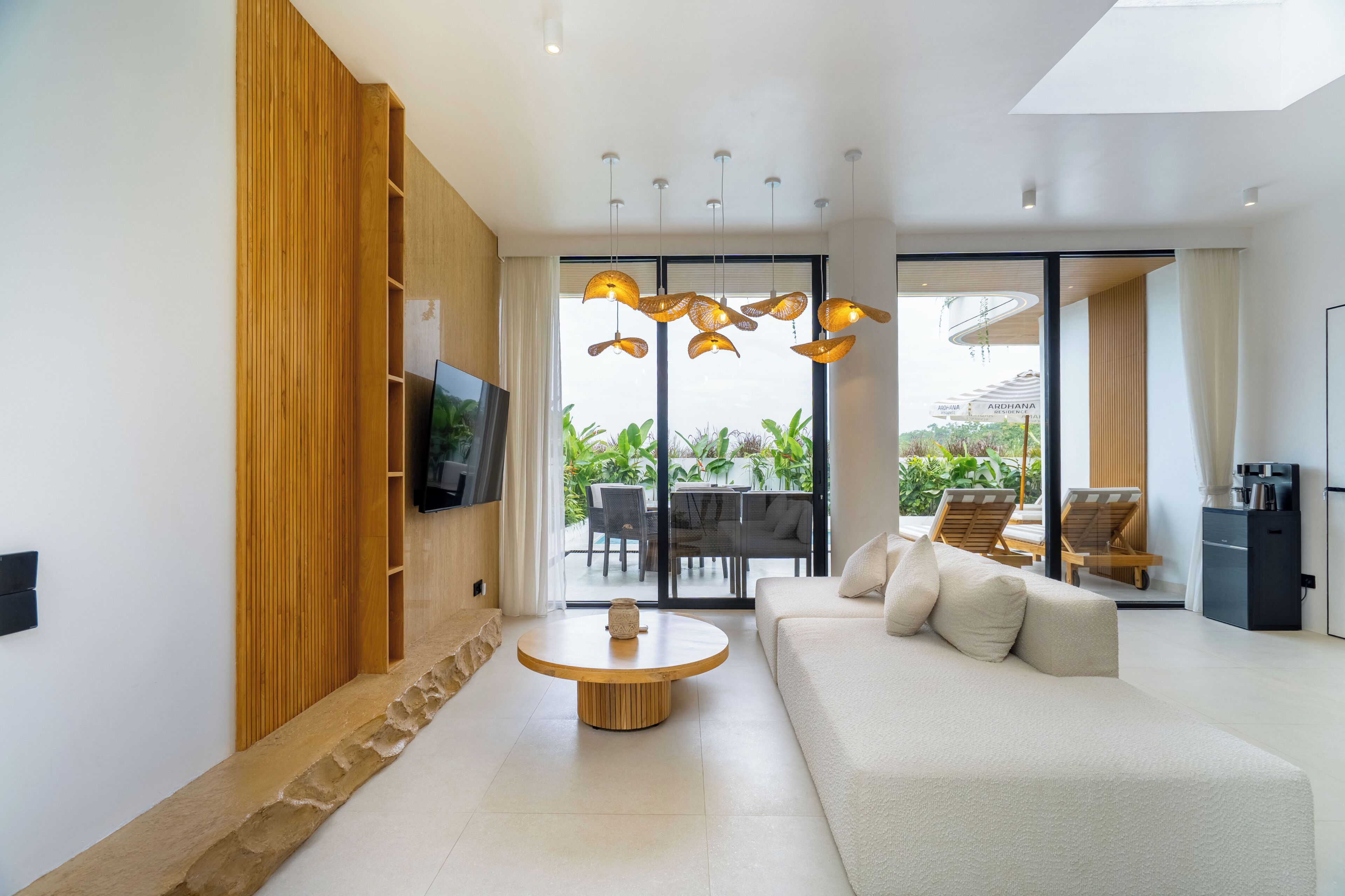 Un salon moderne avec un canapé, une table basse, un téléviseur fixé sur un panneau en bois et une vue sur l’extérieur grâce à de grandes fenêtres.
