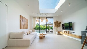 Ein modernes Wohnzimmer mit einer Couch, einem Couchtisch, einem Fernseher und Blick auf einen Garten durch große Fenster.