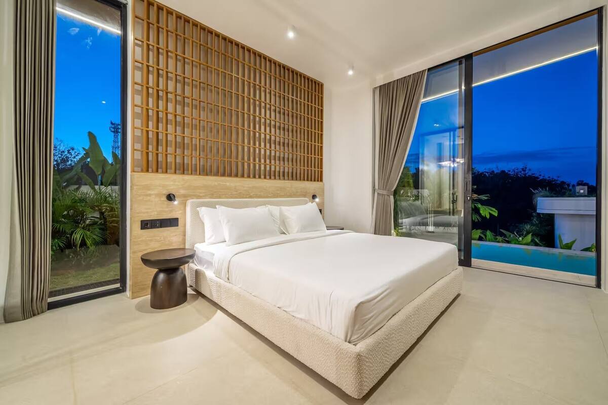 Un dormitorio moderno con una cama grande, cabecera de madera, una mesita de noche pequeña y vistas a una piscina.
