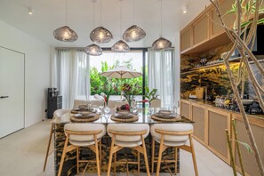 Comedor con mesa de vidrio, cuatro sillas, un paraguero central y lámparas colgantes.
