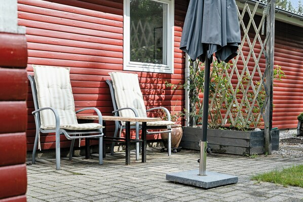 En röd träbyggnad med ett fönster, två stolar, ett bord och ett parasoll.