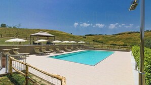Ein Schwimmbecken mit Liegestühlen und Sonnenschirmen, umgeben von einem Zaun und Blick auf Hügel.