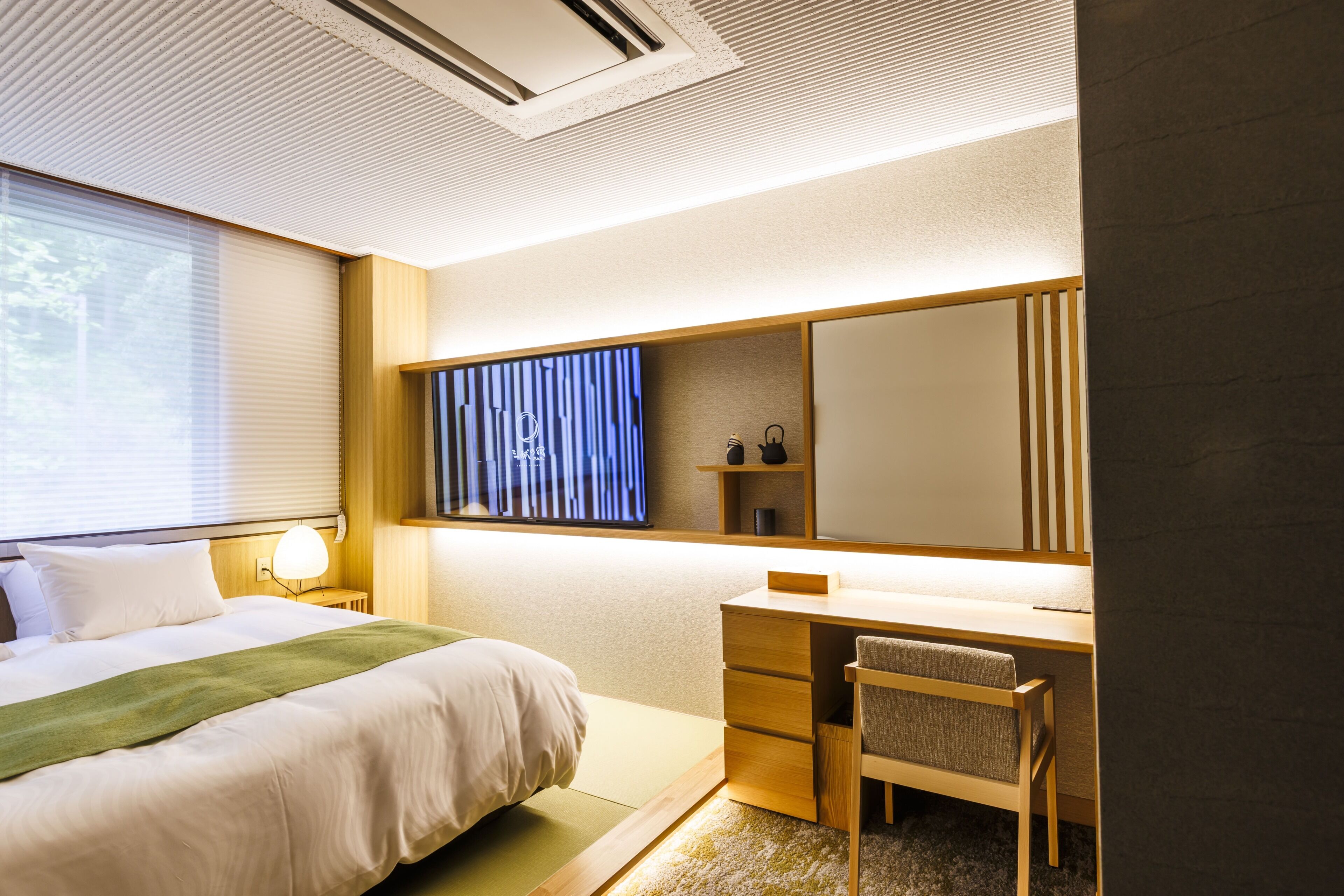 A hotel room with a bed, a desk, a chair, a large mirror, and a window with blinds.