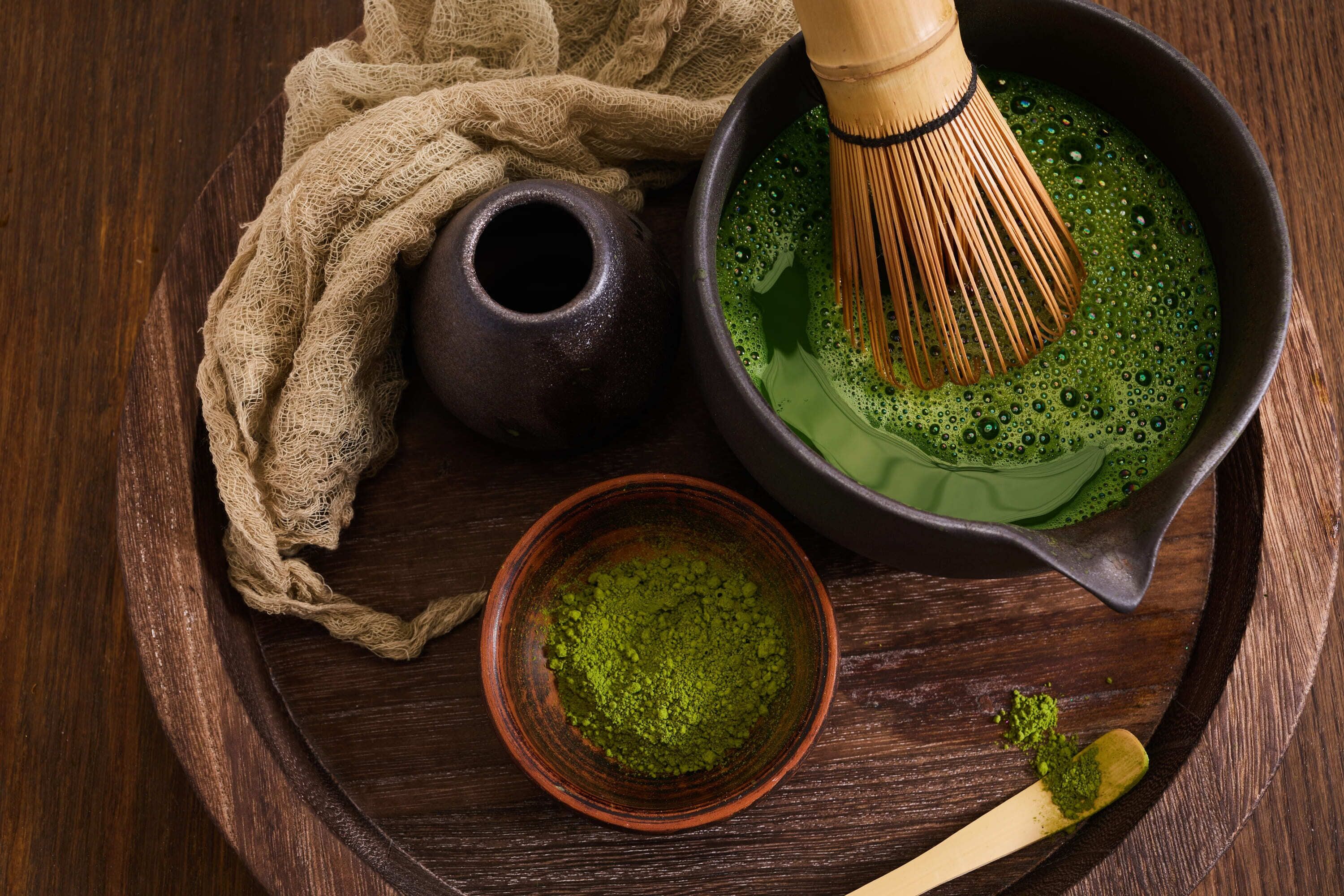 Una bandeja de madera con un bol oscuro que contiene un líquido verde, un batidor, un tazón pequeño de polvo verde y una cuchara de madera.