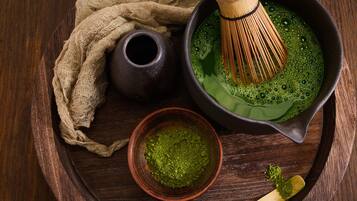 Una bandeja de madera con un bol oscuro que contiene un líquido verde, un batidor, un tazón pequeño de polvo verde y una cuchara de madera.