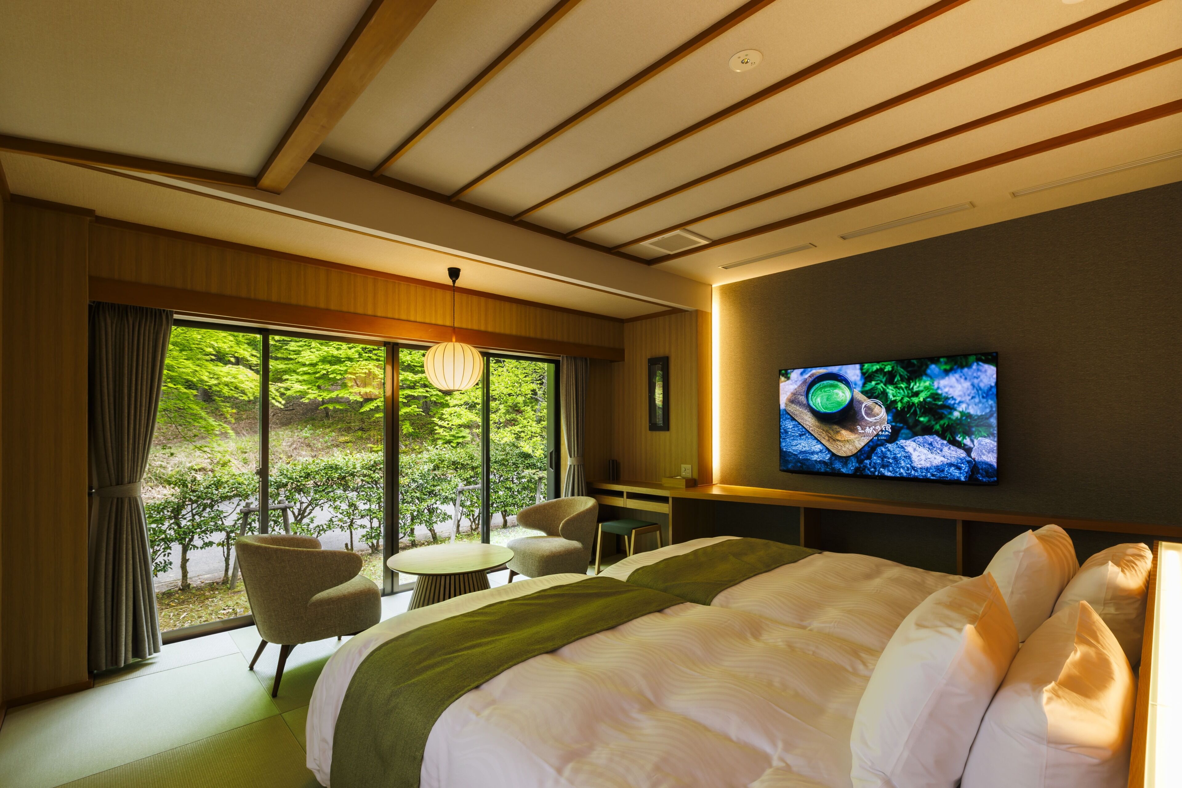 Une chambre d’hôtel équipée d’un lit, d’un bureau, d’une chaise, d’une télévision et offrant une vue sur la verdure par les fenêtres.