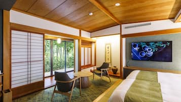 A hotel room with a bed, a TV, a seating area with chairs, a small table, and a view of greenery outside.