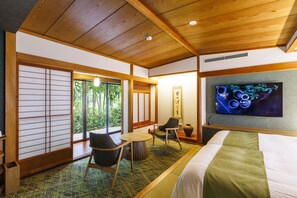 Habitación de hotel con cama, televisor, zona de estar con sillas, una mesita y vistas a zonas verdes.