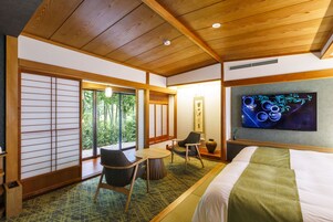 Habitación de hotel con cama, televisor, zona de estar con sillas, una mesita y vistas a zonas verdes.