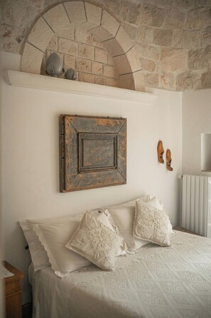 A bedroom with a stone wall, a bed with white bedding, and a rustic wooden frame on the wall. - Historic trullo in Puglia (Ostuni)