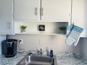 A kitchen with white cabinets, a coffee maker, a sink, a dish soap bottle, and a framed quote on the shelf.