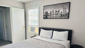 Ett sovrum med en säng, ett fönster med persienner, en tavla med en stadsbild på väggen och en dörr.
