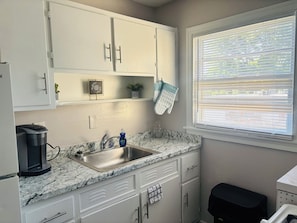 A kitchen with white cabinets, a granite countertop, a coffee maker, a sink, and a window with blinds.