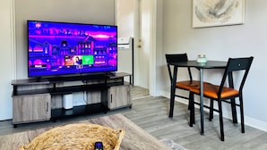 Ett vardagsrum med en platt-TV, ett matbord med stolar, en korg av rotting och en tavla på väggen.