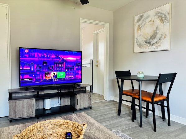 A living room with a flat-screen TV, a dining table with chairs, a wicker basket, and a painting on the wall.