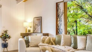 Ein Wohnzimmer mit einer Couch, einem Couchtisch, einer Lampe, einem gerahmten Bild und Blick auf Bäume durch ein Fenster.