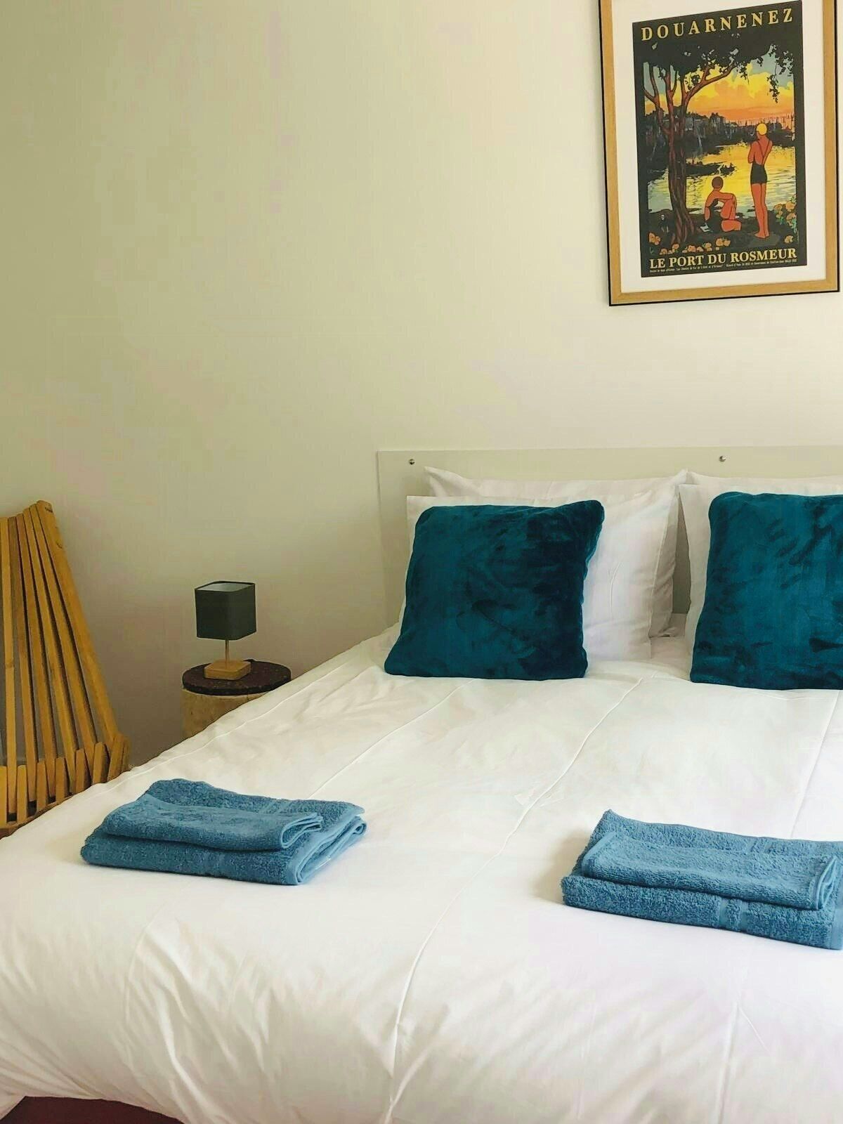 Un lit double avec des oreillers blancs et turquoise, une petite table de chevet avec une lampe et une affiche accrochée au mur.