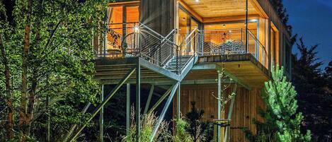 A modern wooden cabin with a glass balcony, surrounded by trees.