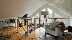 Ein Loft-Zimmer mit schrägverdeckter Decke, Holzbalken, einem weißen Sofa, einer Stehlampe und Blick durch große Fenster.